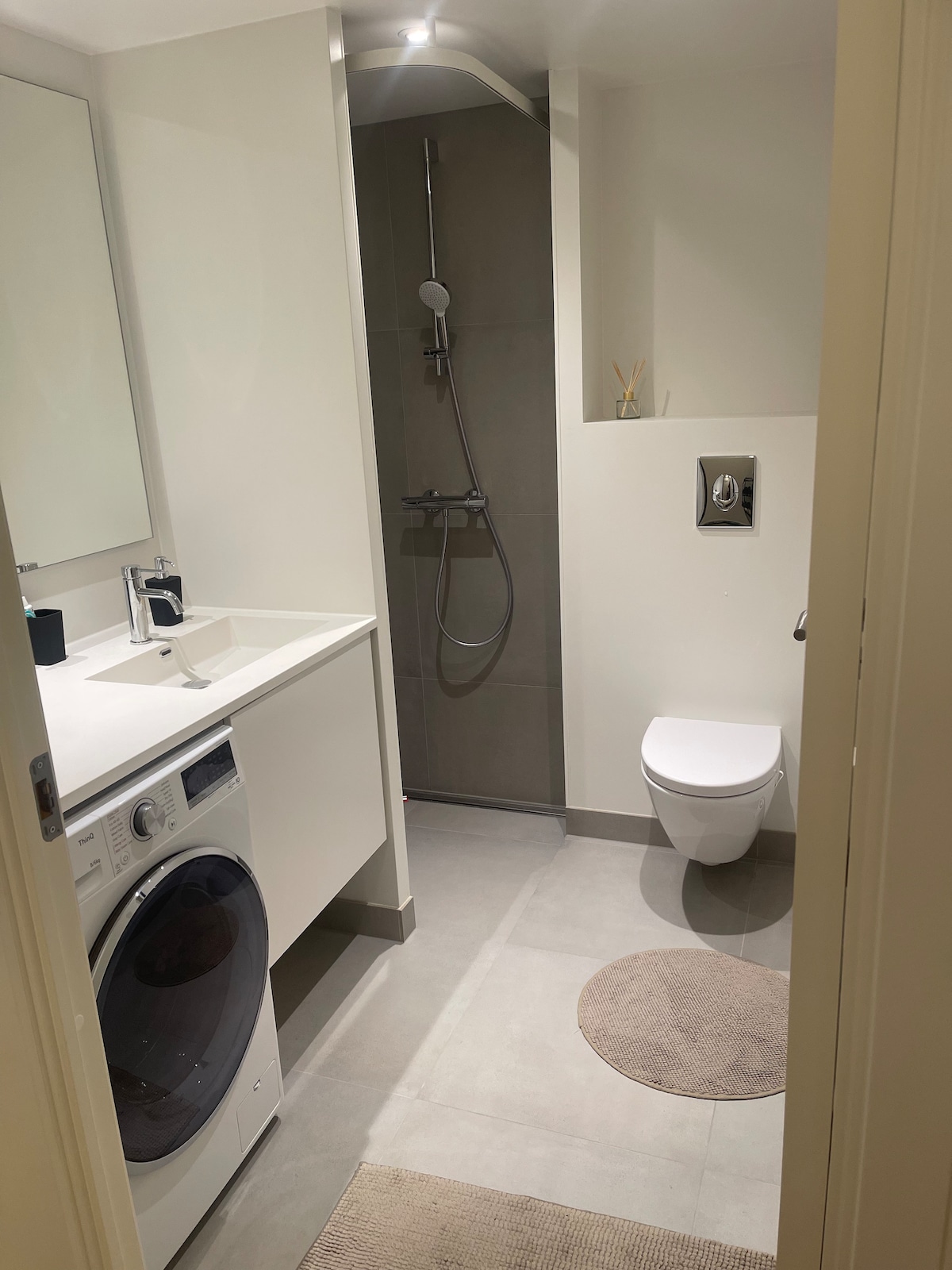 A modern bathroom features a washing machine and a sleek sink countertop. A spacious shower with a glass partition is visible, along with a contemporary toilet. Soft lighting creates a calm environment, while light-colored tiles provide an airy feel.