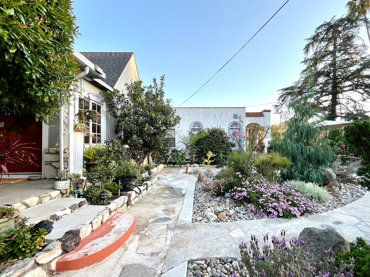A landscaped garden path winds through an array of colorful flowers and plants, leading to a charming Craftsman-style entrance. Stones and pebbles outline the walkway, while lush greenery provides a peaceful ambiance. The clear blue sky complements the serene setting.
