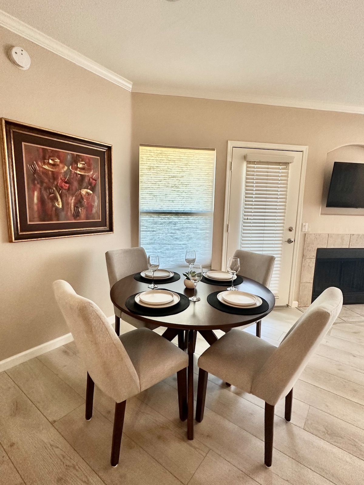 A round dining table is set for four, featuring neutral plates and wine glasses. A window with blinds provides soft natural light, while a framed painting with warm tones adorns the wall. A fireplace is visible in the background, contributing to the space's cozy atmosphere.