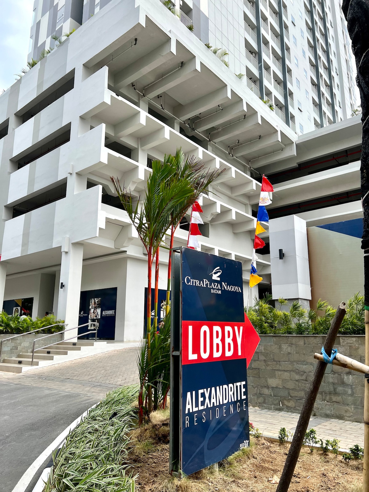 The entrance to CitraPlaza Nagoya is displayed with a clear sign indicating 'LOBBY,' complemented by vibrant flags. Modern architecture and landscaping create a welcoming approach, marked by palm plants along the pathway leading to the lobby entrance.