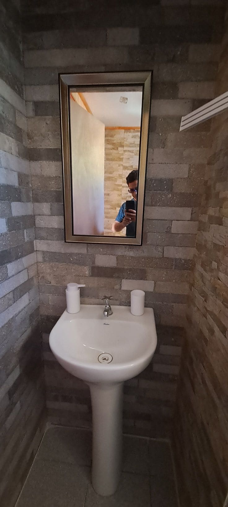 A compact bathroom features a wall-mounted sink with a sleek design, accompanied by two white soap dispensers. The walls are adorned with textured stone tiles, creating a modern atmosphere. A rectangular mirror hangs above the sink, reflecting the natural light from the space.
