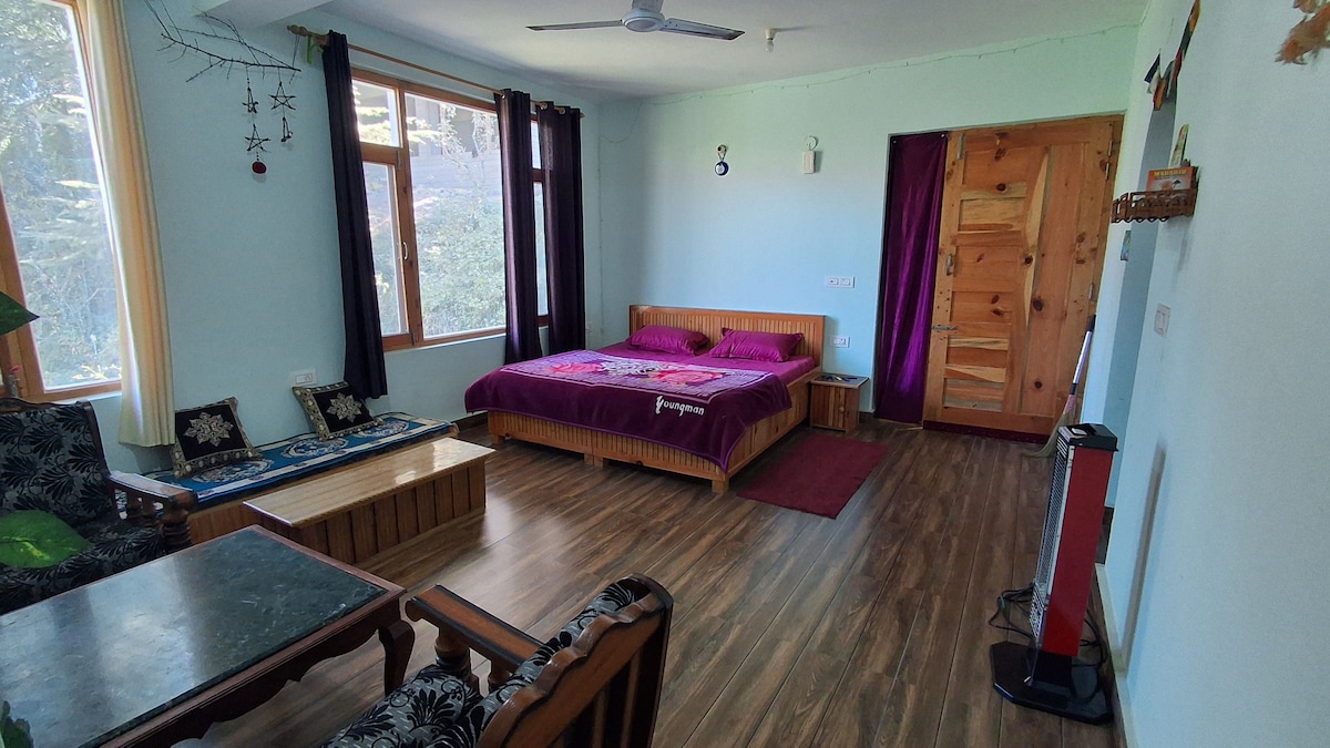 A spacious room is furnished with a large bed covered in deep purple linens. Natural light enters through multiple windows, illuminating the wood accents throughout. A small seating area contains two chairs and a coffee table. A wooden door leads to another area of the homestay.