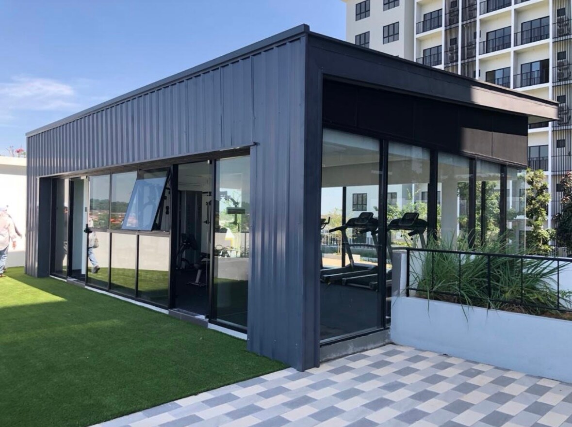 A modern gym facility is featured, with large windows providing visibility of the inside workout area. The exterior showcases a sleek design of dark metal and lush greenery along the pathway. Turf has been laid in front, enhancing the outdoor ambiance.