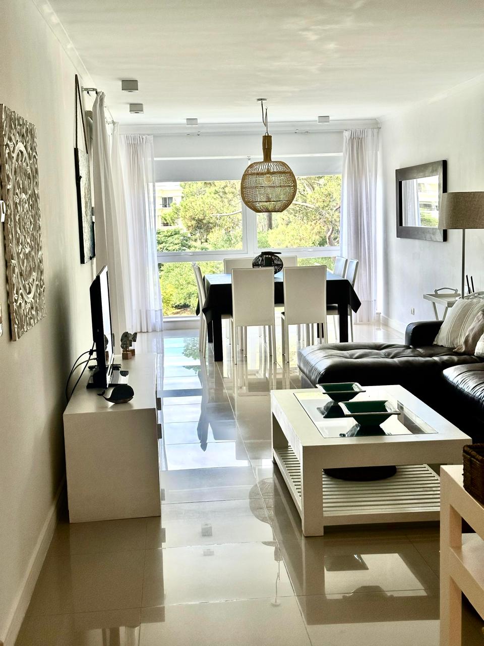 A living area is presented with a black leather sofa, paired with a white coffee table featuring a decorative item. A dining table with white chairs occupies a well-lit space near a large window, offering views of greenery, while modern art adorns the walls.