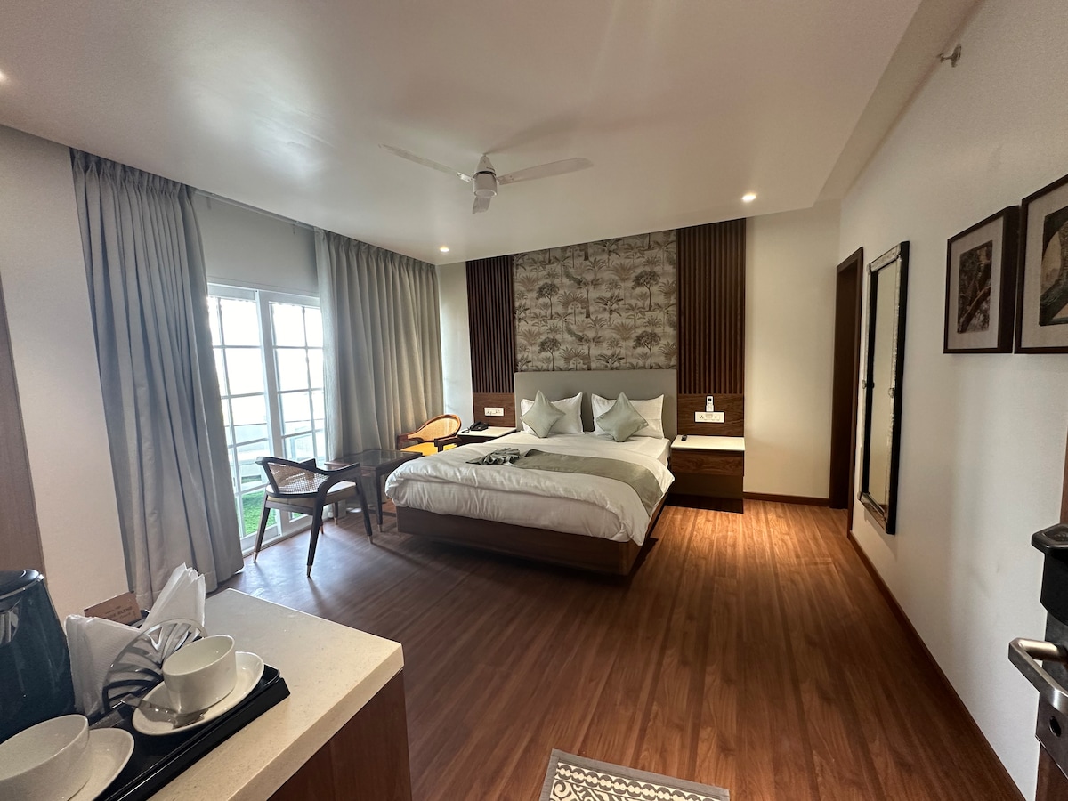 The spacious room features a large bed with multiple pillows, flanked by bedside tables. Natural light pours in through a set of double windows, contrasting with the light wooden flooring. A comfortable chair and a small table are arranged for relaxation, while a kitchenette area is visible.