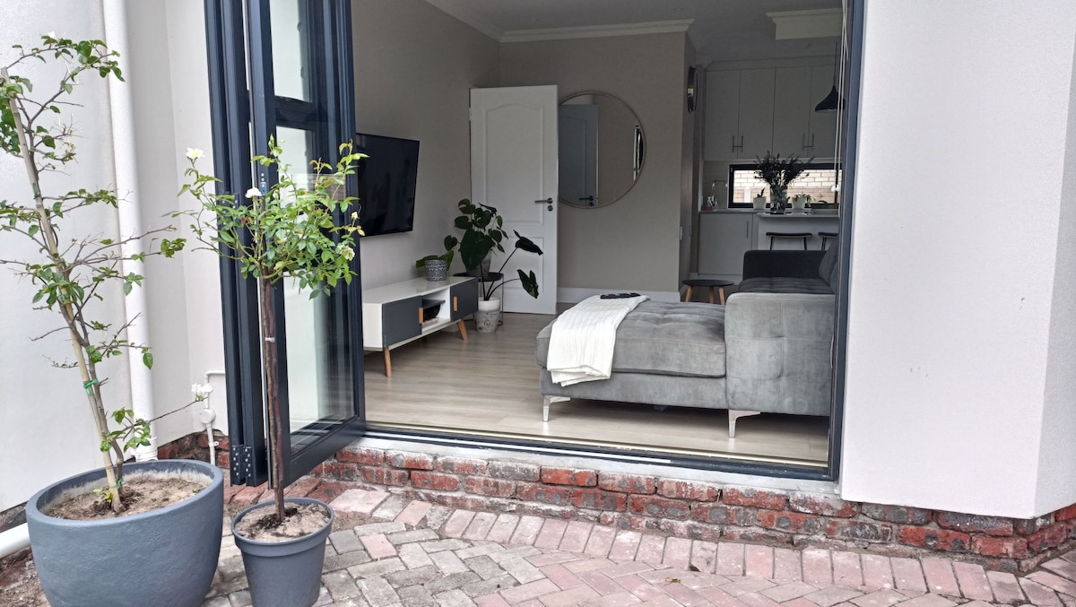 A spacious living area is visible through large glass doors, featuring a comfortable gray sofa and a white media console. A round mirror reflects light, and potted plants add a touch of greenery. A cozy throw is casually draped over the sofa.
