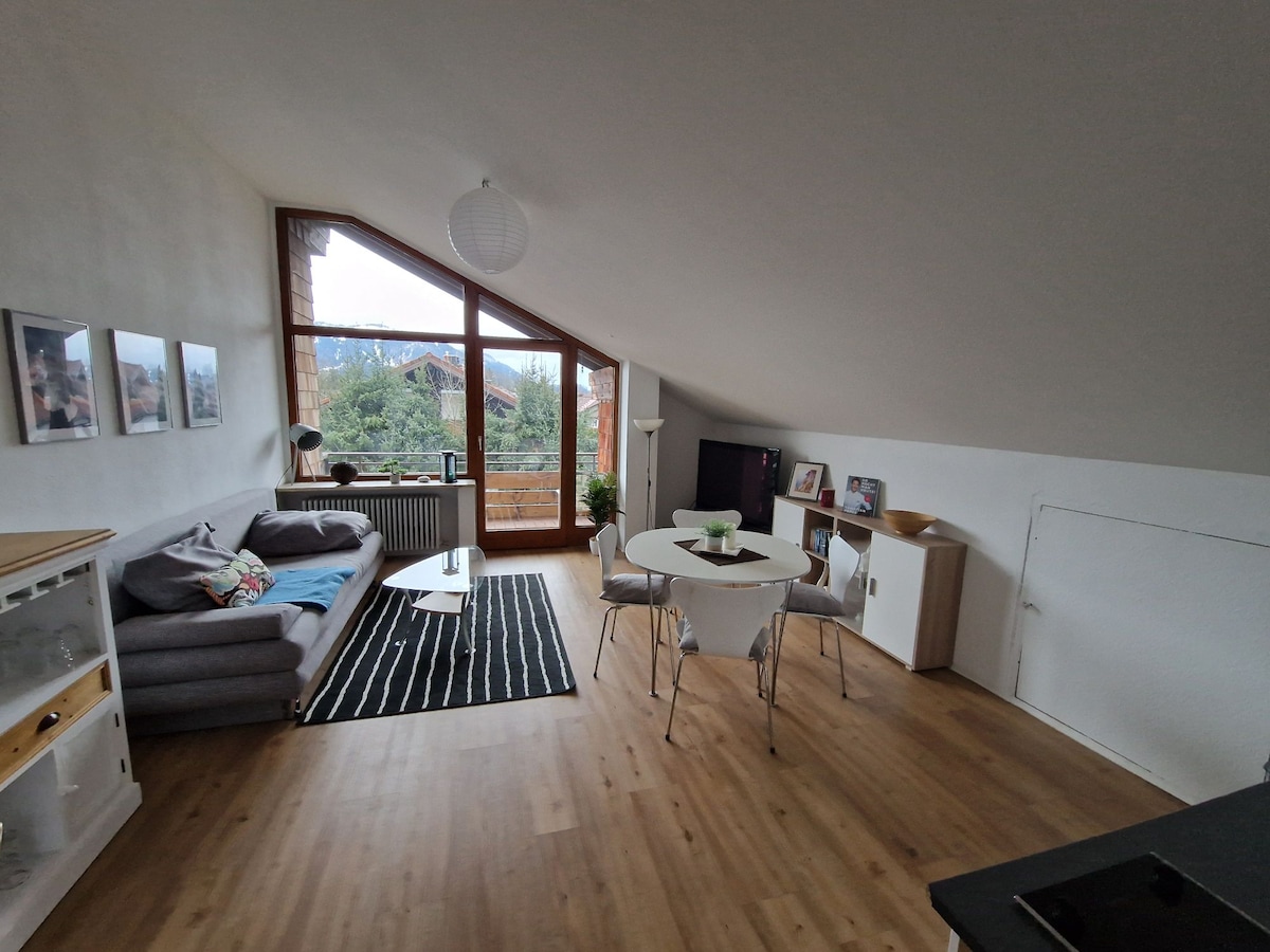 A spacious living area is illuminated by natural light from large triangular windows. A gray sofa is positioned alongside a striped rug, while a round dining table with white chairs is centrally located. Decorative elements include a media unit and wall-mounted artwork.