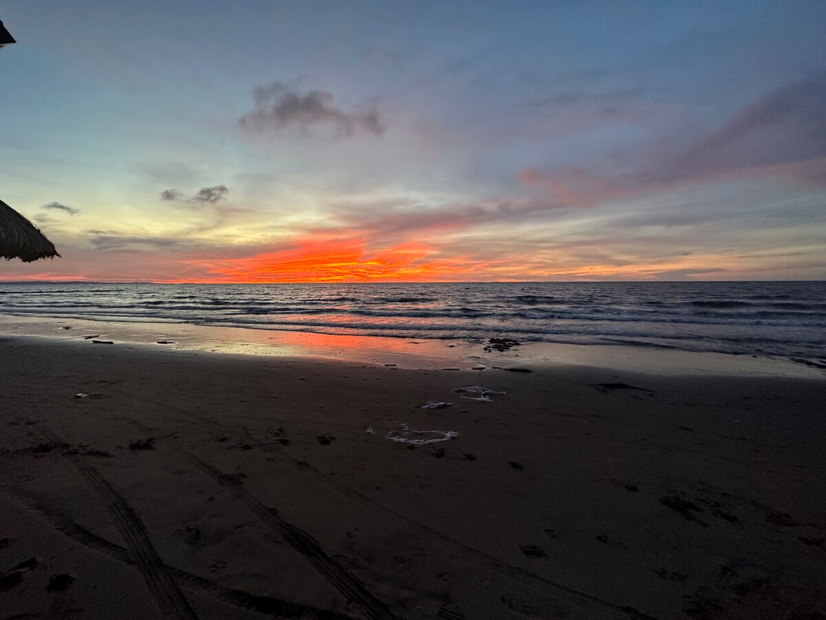 A serene beach scene captures a vibrant sunset, with hues of orange, pink, and purple reflected on the water. Gentle waves lap at the shore, and smooth sand stretches along the coastline, creating a peaceful atmosphere.