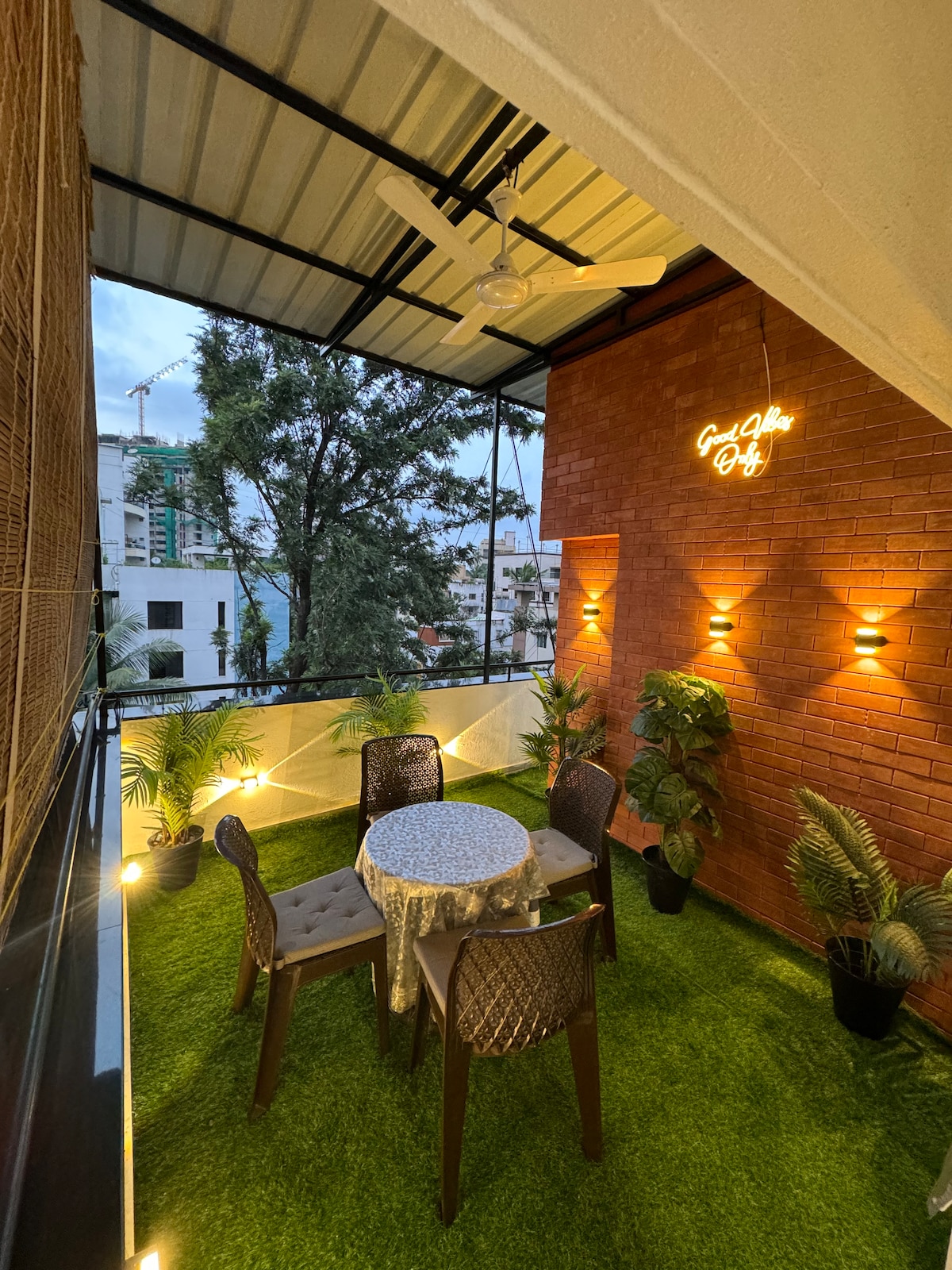 A charming balcony area features a round table covered with a tablecloth, surrounded by four chairs. Lush greenery is integrated into the decor, with potted plants placed at intervals. Soft lighting enhances the space, creating a relaxed atmosphere for outdoor enjoyment.