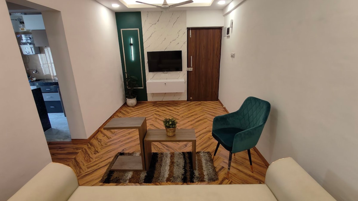 A modern living area is highlighted by a rich wooden herringbone floor. A green accent chair is positioned next to a wooden coffee table, which holds a small potted plant. A neutral rug lies beneath, and a mounted TV is visible on the opposite wall.