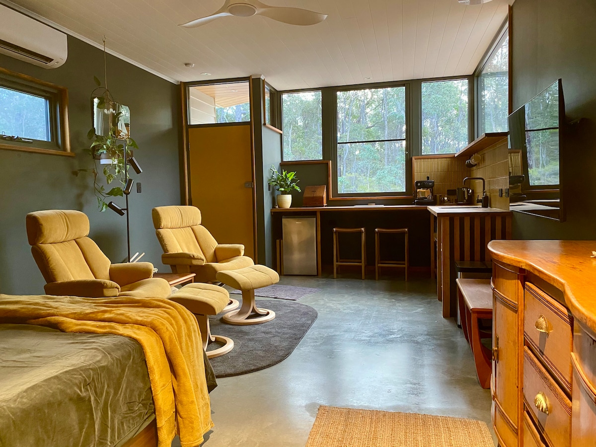 An open living space features large windows overlooking the forest. Two comfortable armchairs are set beside a modern kitchen area, which includes appliances like a kettle and espresso machine. A cozy bed is draped with a soft blanket, enhancing the calm ambiance.