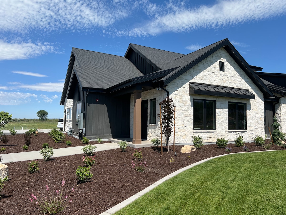 The exterior of the home features a combination of stone and dark siding, complemented by a well-maintained lawn and landscaped flower beds. Large windows provide natural light, and a covered entryway leads into the house. A clear blue sky adds to the serene atmosphere.