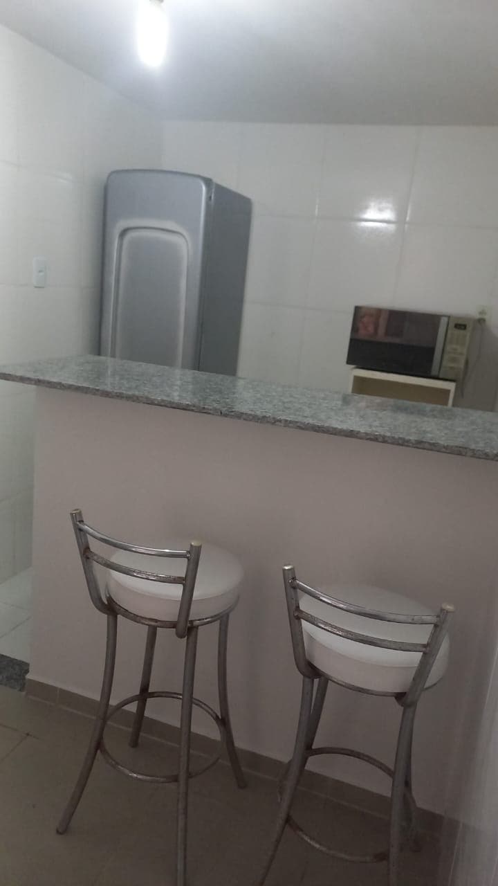A modern kitchen is presented with sleek white tiles and a granite countertop. Two high stools are positioned at the counter. A stainless steel refrigerator and a microwave are visible, providing essential appliances for meal preparation.