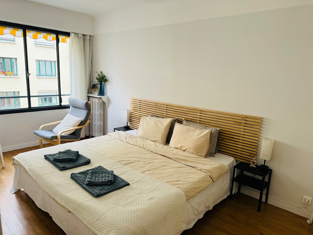 A spacious bedroom features a king-size bed with neutral bedding. Soft pillows adorn the headboard. A comfortable armchair sits beside the bed, while a small nightstand holds lamps. Light enters through the large window, enhancing the calming atmosphere.