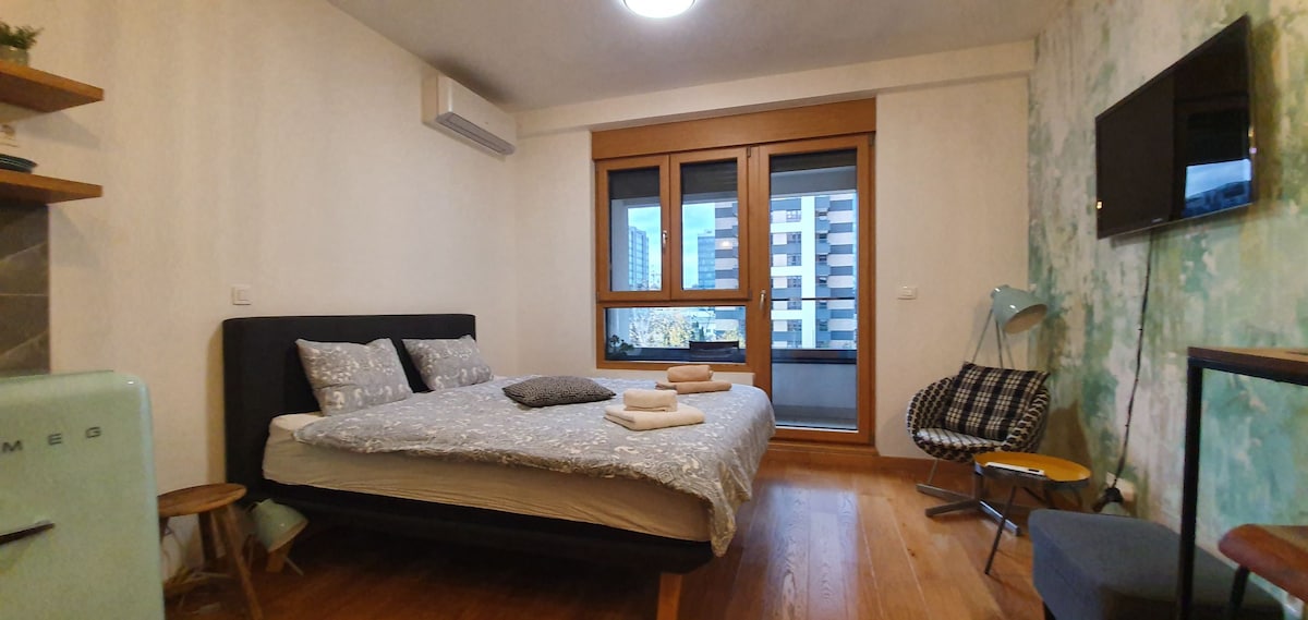 A modern bedroom features a spacious bed dressed with light-colored linens and neatly arranged towels. Large windows provide natural light and views of the surrounding area. A small armchair and a TV are positioned for convenience, complemented by wooden flooring and a subtle green accent wall.