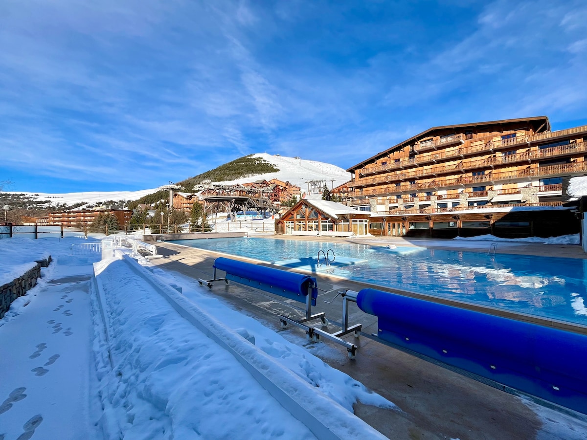 An outdoor swimming pool is visible, surrounded by snow-covered ground. Multiple lounge chairs line the edge, while the expansive mountains loom in the background. Buildings with multiple levels are seen, showcasing a mix of architectural styles under a clear blue sky.