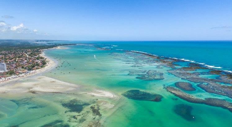 An expansive coastal view reveals a blend of sandy beaches and turquoise waters. Rocky formations extend into the sea, with a clear sky above and a few boats scattered on the horizon. The vibrant colors of the water contrast with the sandy shorelines.