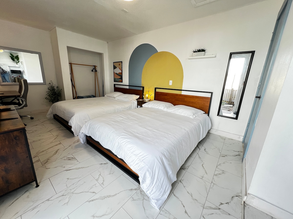 The studio features two spacious beds, one queen and one full, positioned against a modern accent wall in soft colors. A minimalist desk and chair provide a dedicated work space, while natural light filters through a nearby window, adding to the bright atmosphere.