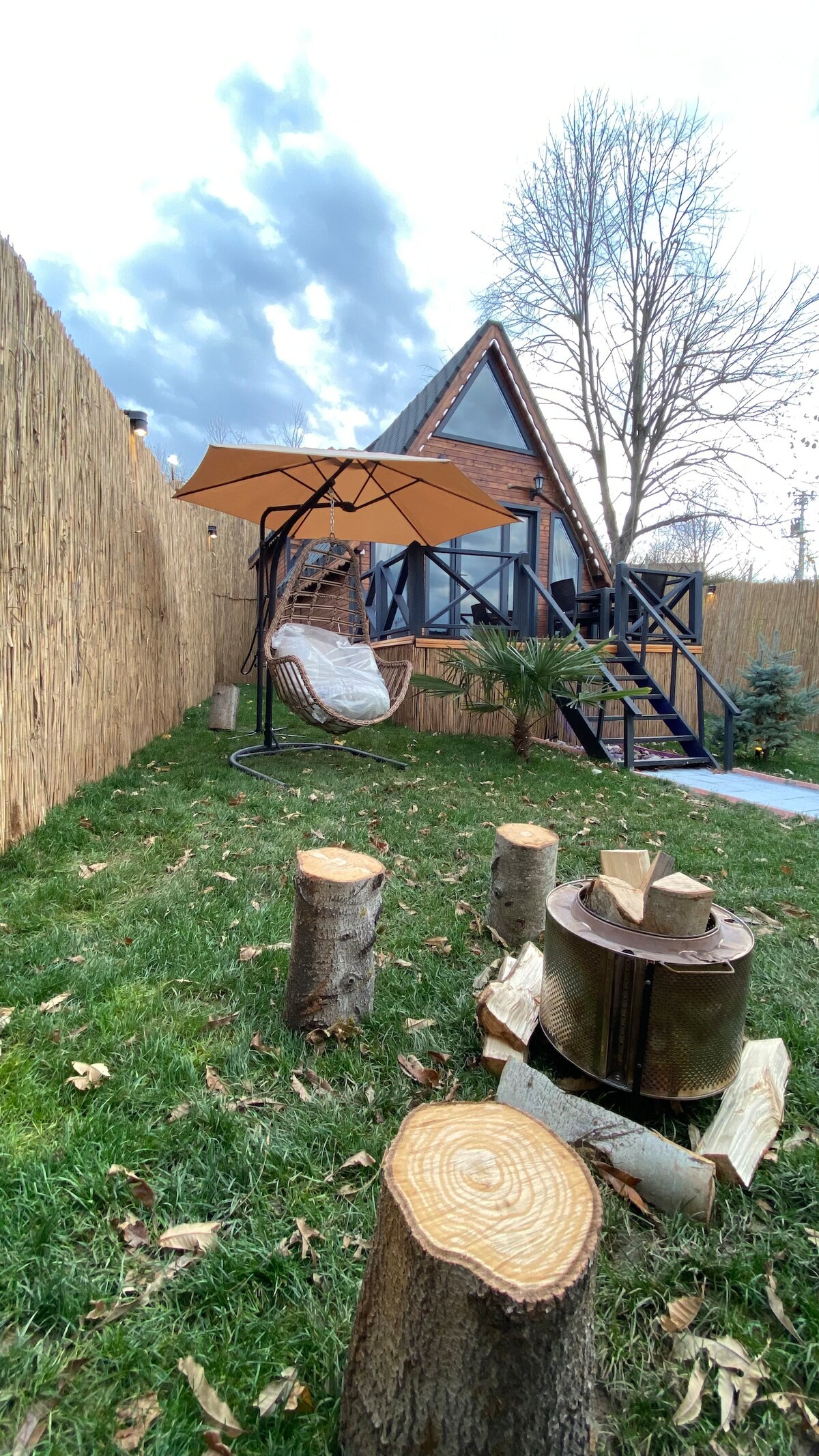 A charming outdoor area features a hanging chair beneath an umbrella, surrounded by lush green grass. Cut tree stumps create a rustic, natural border around a fire pit. A modern A-frame house is visible in the background, framed by tall grass fencing and trees.