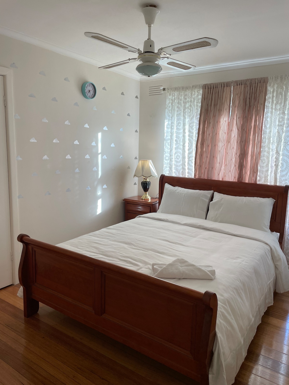 A well-lit bedroom features a spacious bed with crisp white linens. A wooden nightstand is positioned beside the bed, holding a lamp. Soft curtains frame the window, and light blue cloud decals are displayed on the wall, complemented by a wall clock.