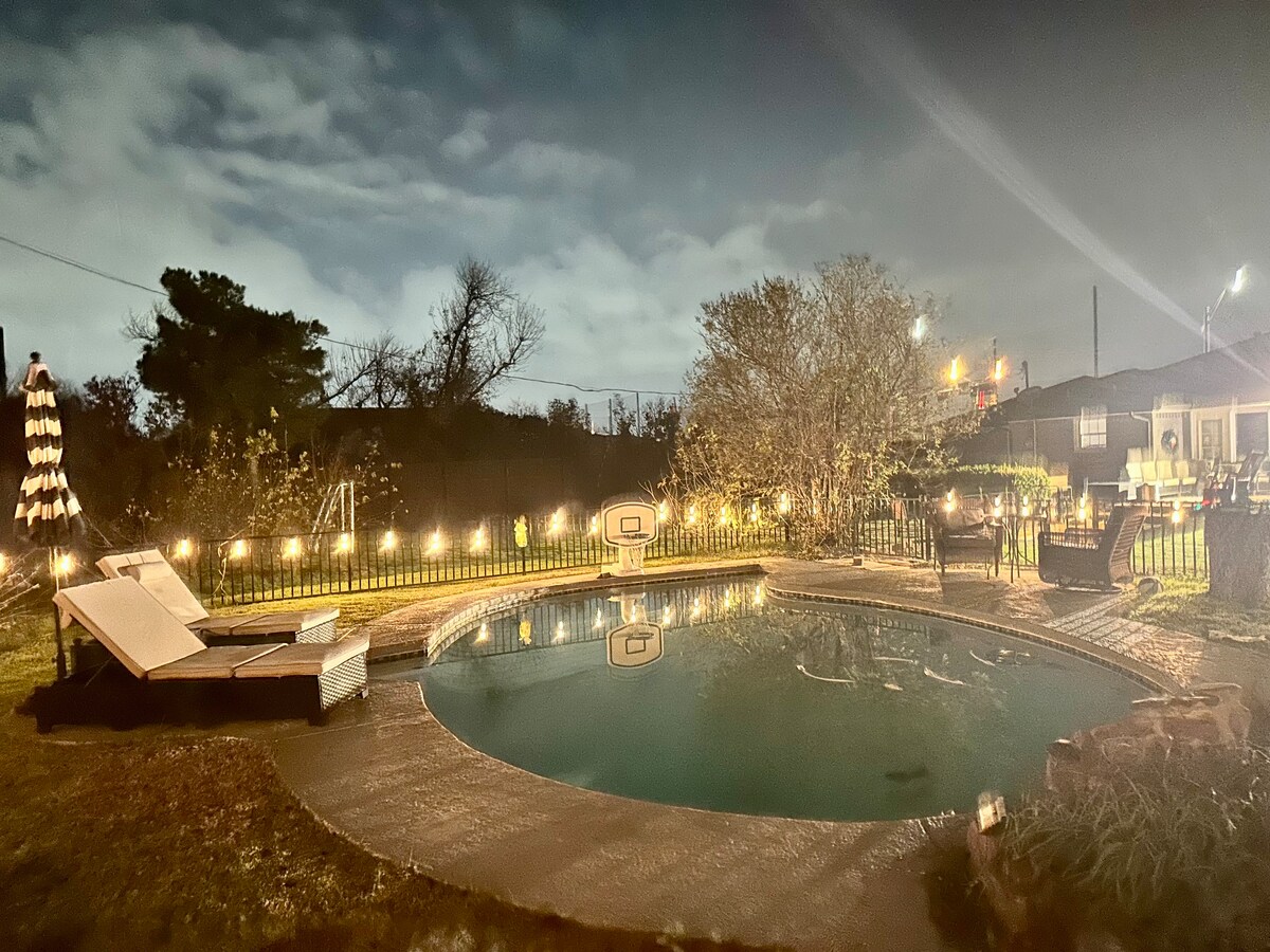 A tranquil pool area is showcased under a night sky, featuring a circular swimming pool with gently rippling water. Lounge chairs are positioned nearby, illuminated by soft lights strung along the fence. A cozy seating arrangement is visible, enhancing the inviting outdoor space.