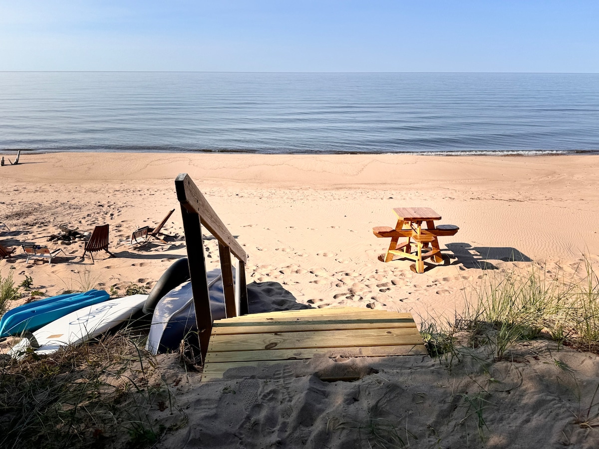 A wooden staircase leads down to a wide expanse of sand beach, with gentle waves lapping at the shore. A picnic table sits on the sand, accompanied by lounge chairs arranged nearby. The tranquil water reflects the clear blue sky, creating a serene atmosphere.