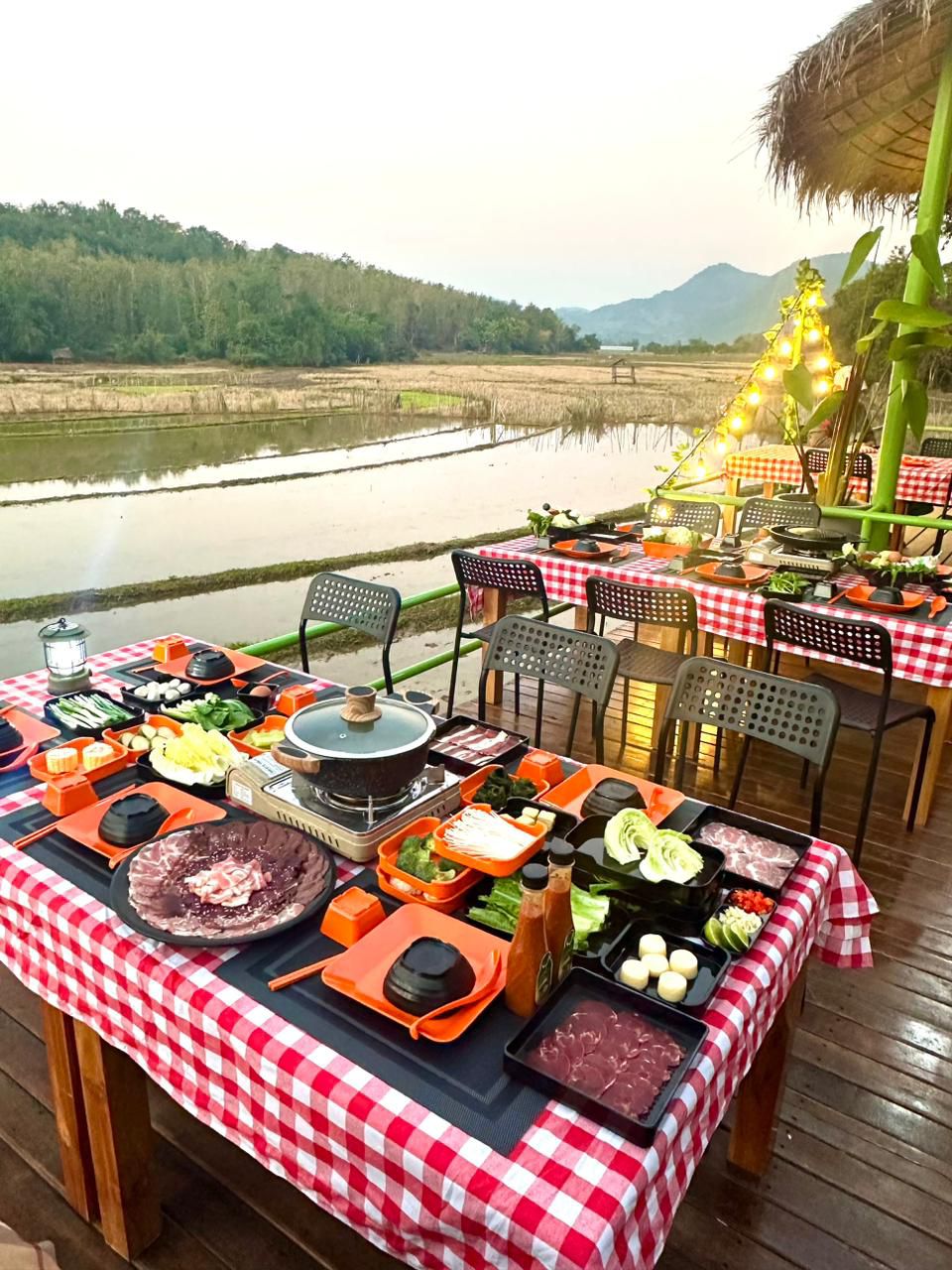 An outdoor dining area features several tables covered with checkered tablecloths, set with a variety of colorful dishes and fresh ingredients. A serene view of nearby rice paddies and distant hills can be observed in the background, illuminated by soft lighting from surrounding trees.