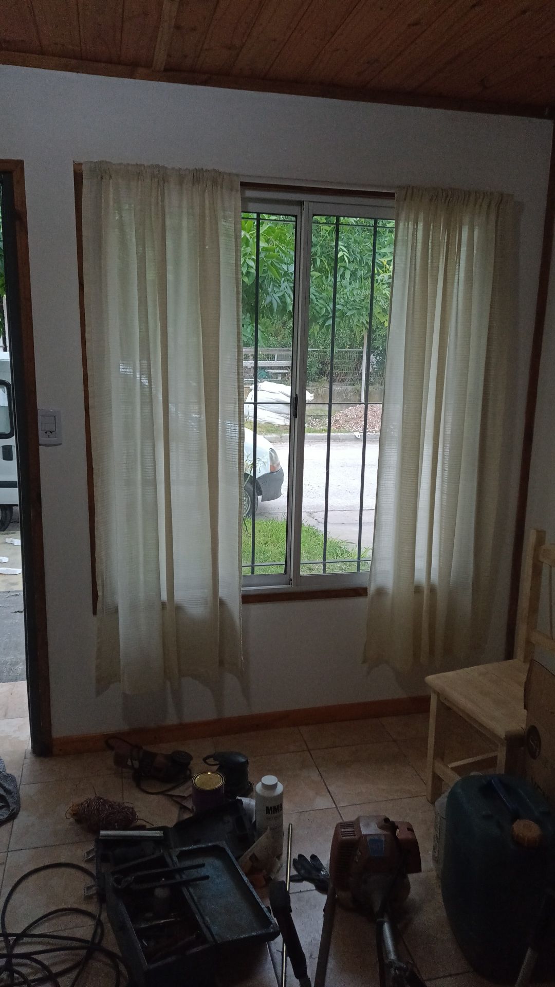A window with sheer curtains is positioned on a white wall, allowing natural light to filter through. Outside, greenery is visible, while various tools and containers are arranged on the floor, giving a glimpse of an ongoing project within the space.