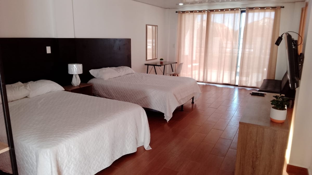 A spacious room features two neatly made beds with white bedding. Large windows allow natural light to fill the space, complemented by light curtains. A television and a small table with a chair are visible, alongside a minimal decorative plant on the dresser.