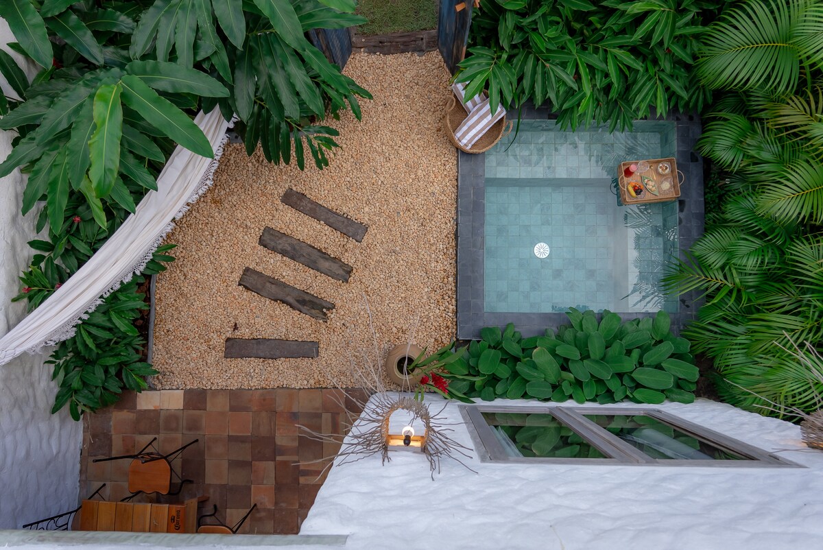 An aerial view captures a tranquil private garden, featuring a small pool surrounded by lush greenery and pebbles. Wooden planks are arranged in a casual manner, complemented by a relaxing seating area with a small table.