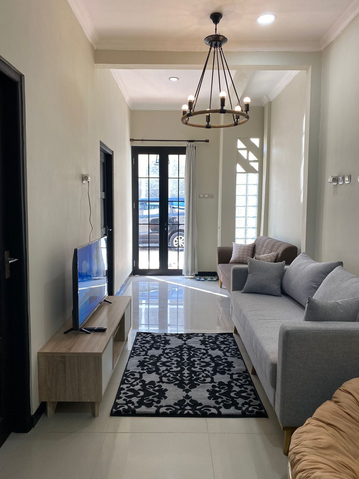 An inviting living area features a comfortable gray sofa and a neutral armchair, complemented by a patterned area rug. A wooden TV stand holds a flat-screen television, while large windows provide natural light and a view of the outdoors.