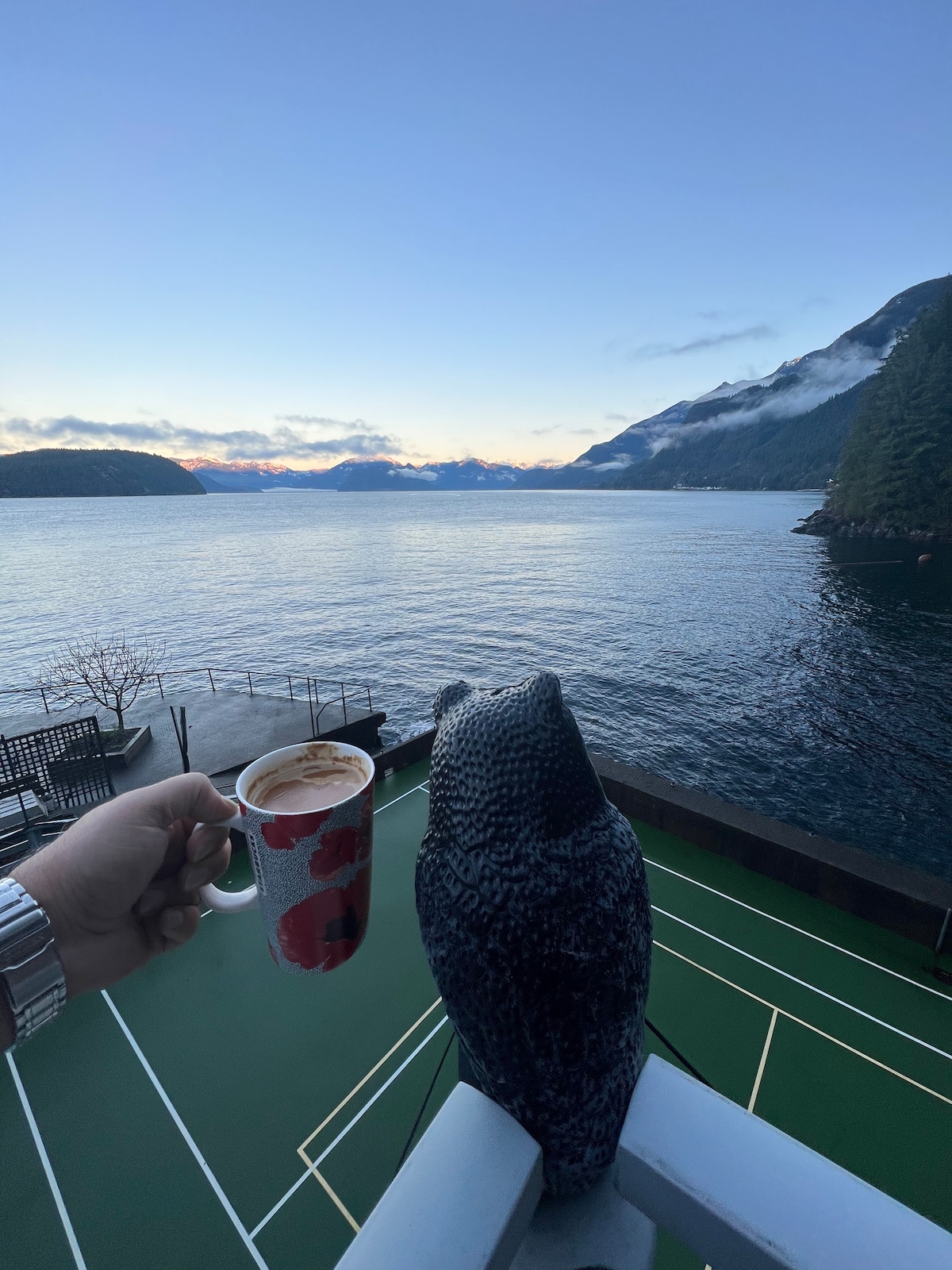 A hand holds a coffee cup adorned with red flowers, positioned beside a decorative owl statue on a balcony railing. The view showcases calm waters reflecting the sky, with mountains in the background and clouds hovering above.