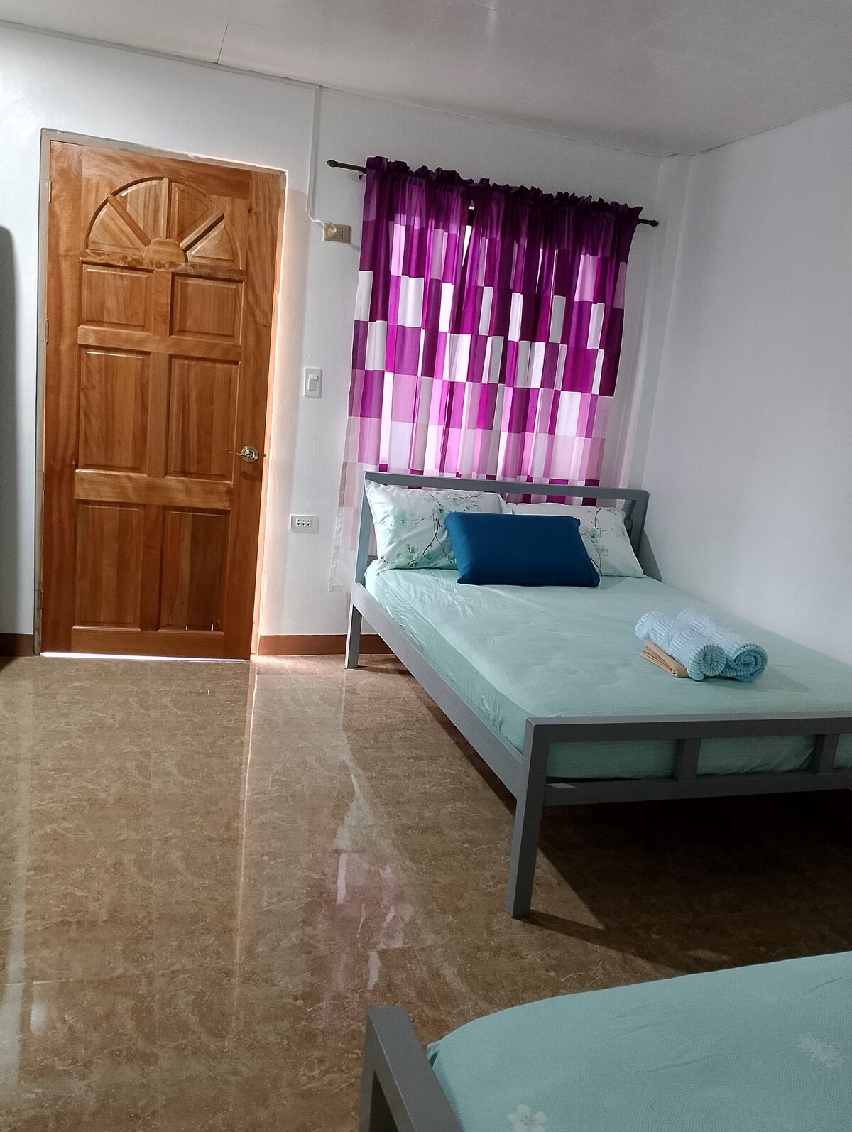 A well-lit room features two spacious beds with light bedding. A decorative blue pillow and rolled towels are placed on one bed. A door with a rounded top is visible, alongside a window dressed with purple and white patterned curtains.