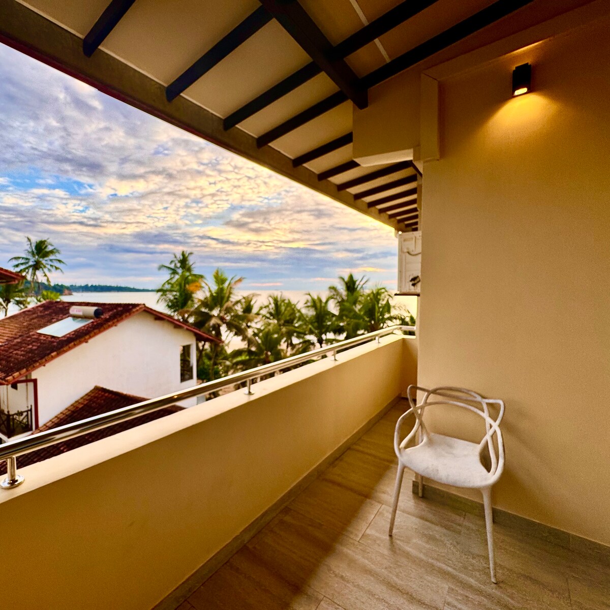 A private terrace is featured, showcasing a white chair and a clear view of the surrounding palm trees and ocean. The soft colors of the sky are captured as the sun rises or sets, adding to the serene ambiance of the space.
