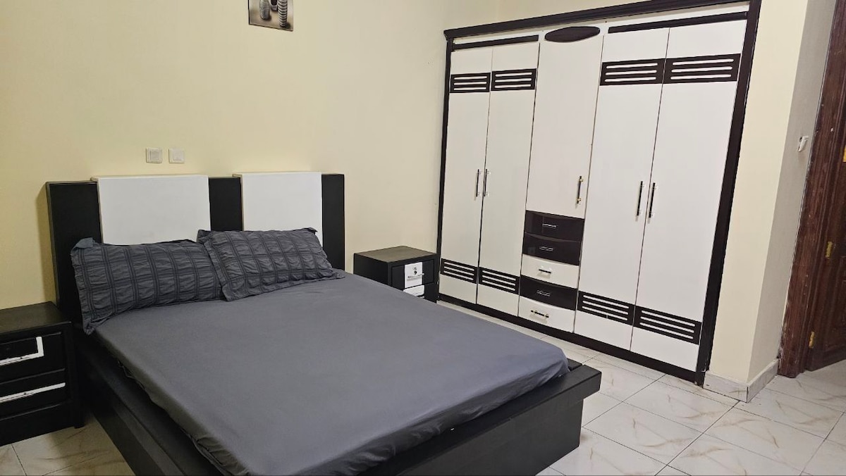 A sleeping area features a spacious black bed with gray linens and decorative pillows. A matching nightstand is positioned beside the bed. A large wardrobe with black and white detailing stands against a light-colored wall, offering ample storage space.