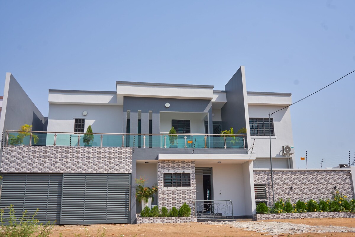 A modern duplex home is displayed, featuring a sleek exterior with a mix of textured white and patterned stone walls. A covered balcony adorned with greenery overlooks the entrance. The front yard is neatly landscaped, showcasing manicured plants against a sunny blue sky.
