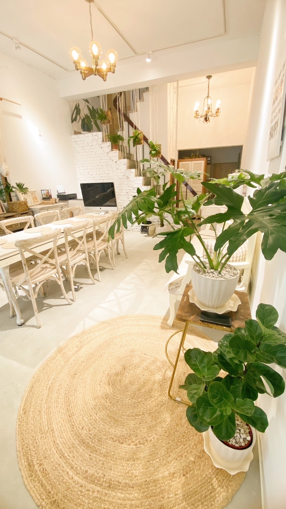 A spacious dining area features a row of wooden chairs surrounding a long table. A decorative rug lies on the floor, while plants add greenery to the space. Stairs lead to the upper level, with warm light illuminating the area from a ceiling chandelier.