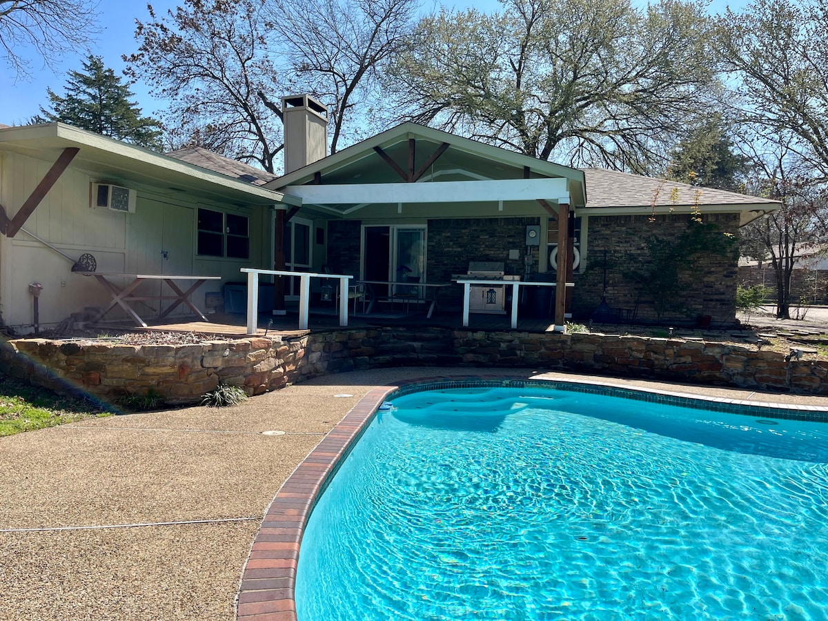 A spacious outdoor area is presented, featuring a clear blue pool adjacent to a stone patio. The structure showcases a covered porch with seating options and a gabled roof, surrounded by trees that provide partial shade.