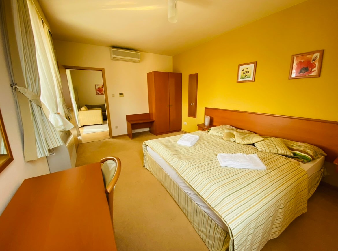 A bright bedroom features a spacious bed covered with a striped blanket. Light-colored walls are complemented by framed artwork. A work desk and a wooden cabinet provide functional space, while a window invites natural light. A mirror reflects the room's layout.