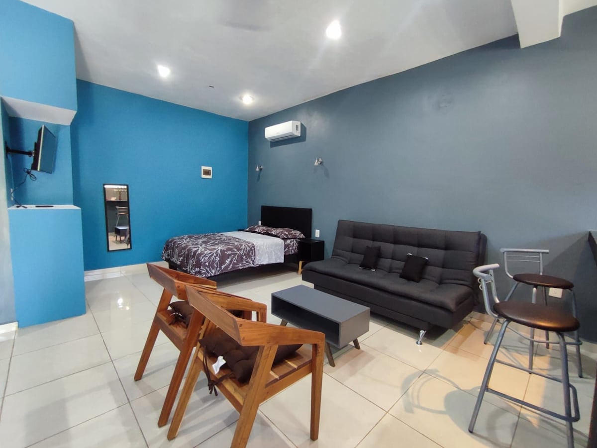 A modern living space is showcased, featuring a dark couch and a spacious bed against a teal wall. Simple wooden chairs and a small table create a functional seating area, while a flat-screen TV is mounted on one wall, enhancing the open layout.