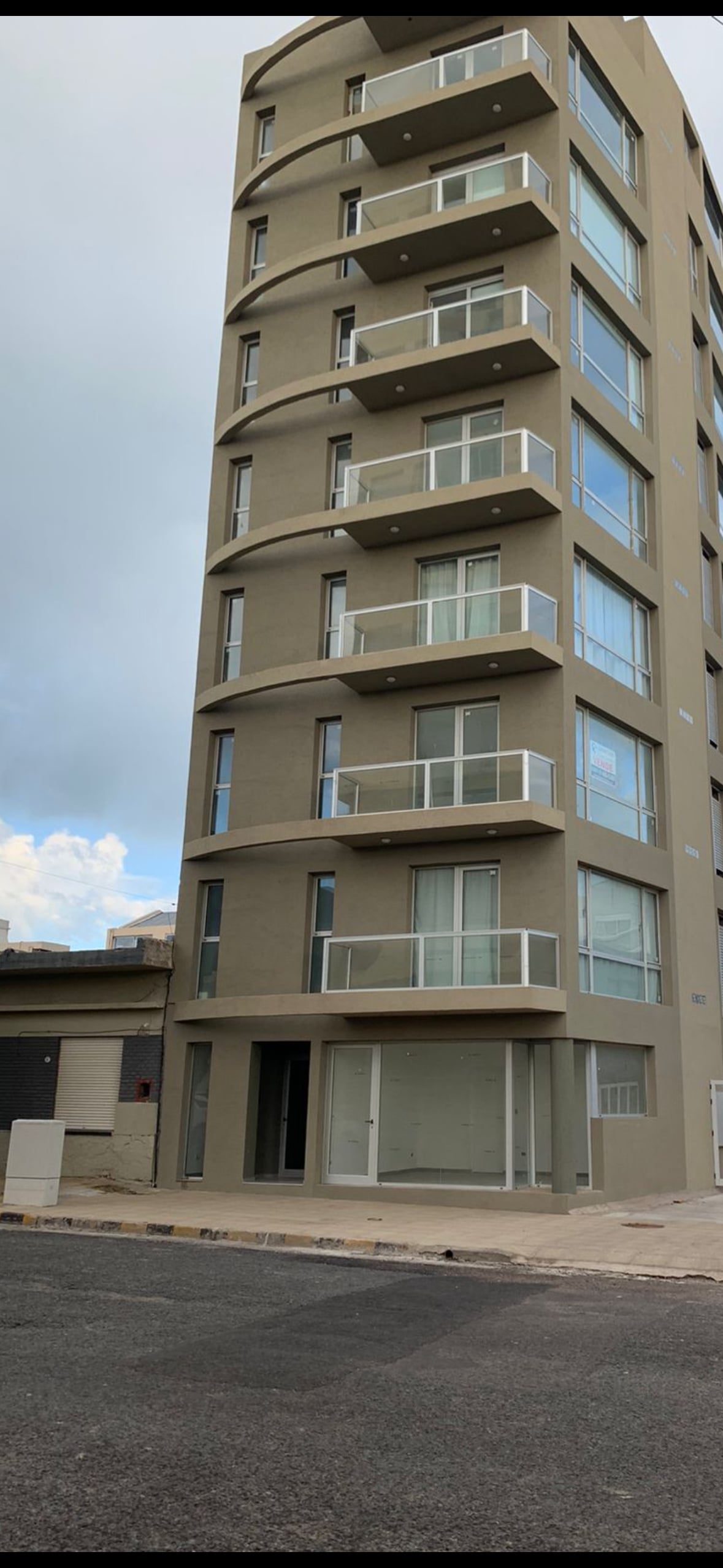 A modern six-story building is shown, featuring a minimalist design with large glass balconies on each level. The facade is a neutral color, and the ground floor displays large windows, providing ample natural light. The entrance is located at street level.