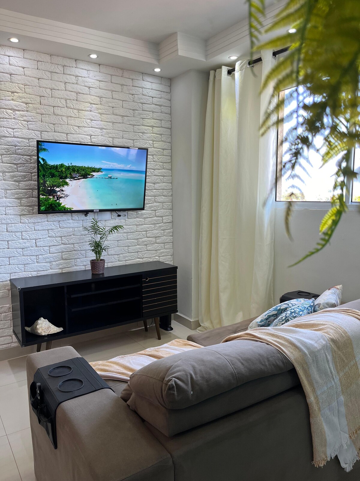 A comfortable living area features a soft gray sofa positioned opposite a wall-mounted television, displaying a serene beach scene. Natural light enters through two bright windows adorned with sheer curtains, enhancing the space's openness. A small potted plant adds a touch of greenery.