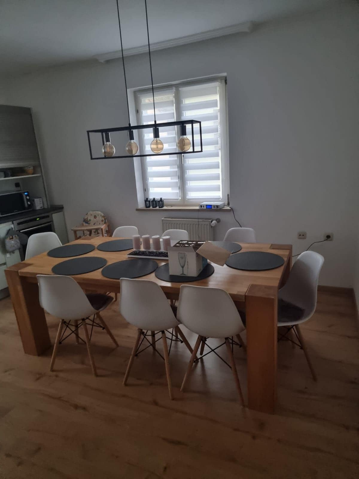 A dining area features a large wooden table surrounded by six white chairs. Above the table, a black pendant light with four glass bulbs illuminates the space. Natural light enters through a window with horizontal blinds, creating a warm and inviting atmosphere.