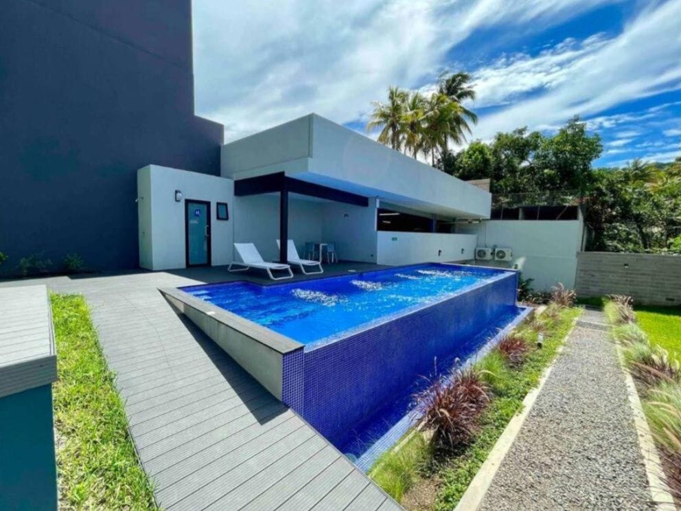 A modern outdoor space features a striking infinity pool surrounded by wooden decking. Lounge chairs offer a place to relax, with vibrant greenery visible in the background. The area is bright, complemented by a clear blue sky and lush tropical plants.