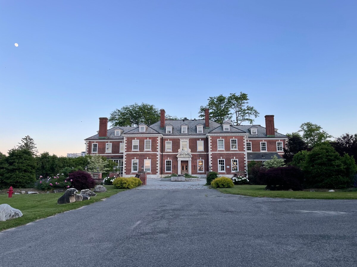 A grand estate is framed in a tranquil setting, featuring a symmetrical brick facade. Lush greenery surrounds the property, enhancing its stately presence. The clear sky exhibits a hint of moonlight, creating a serene backdrop for this magnificent structure.