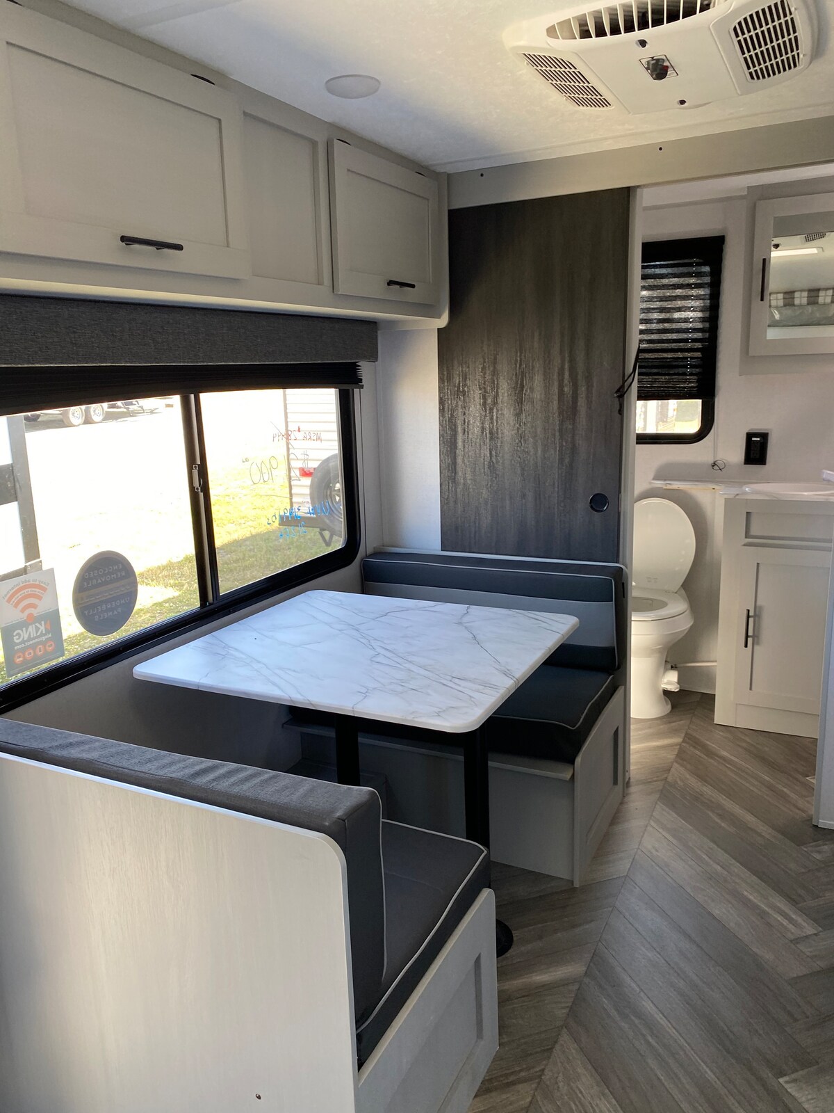 A cozy interior of the camper showcases a small dining area with a marble-topped table surrounded by cushioned seating. A window allows natural light to enter the space, providing a view of the outdoors. A compact bathroom is visible in the background.