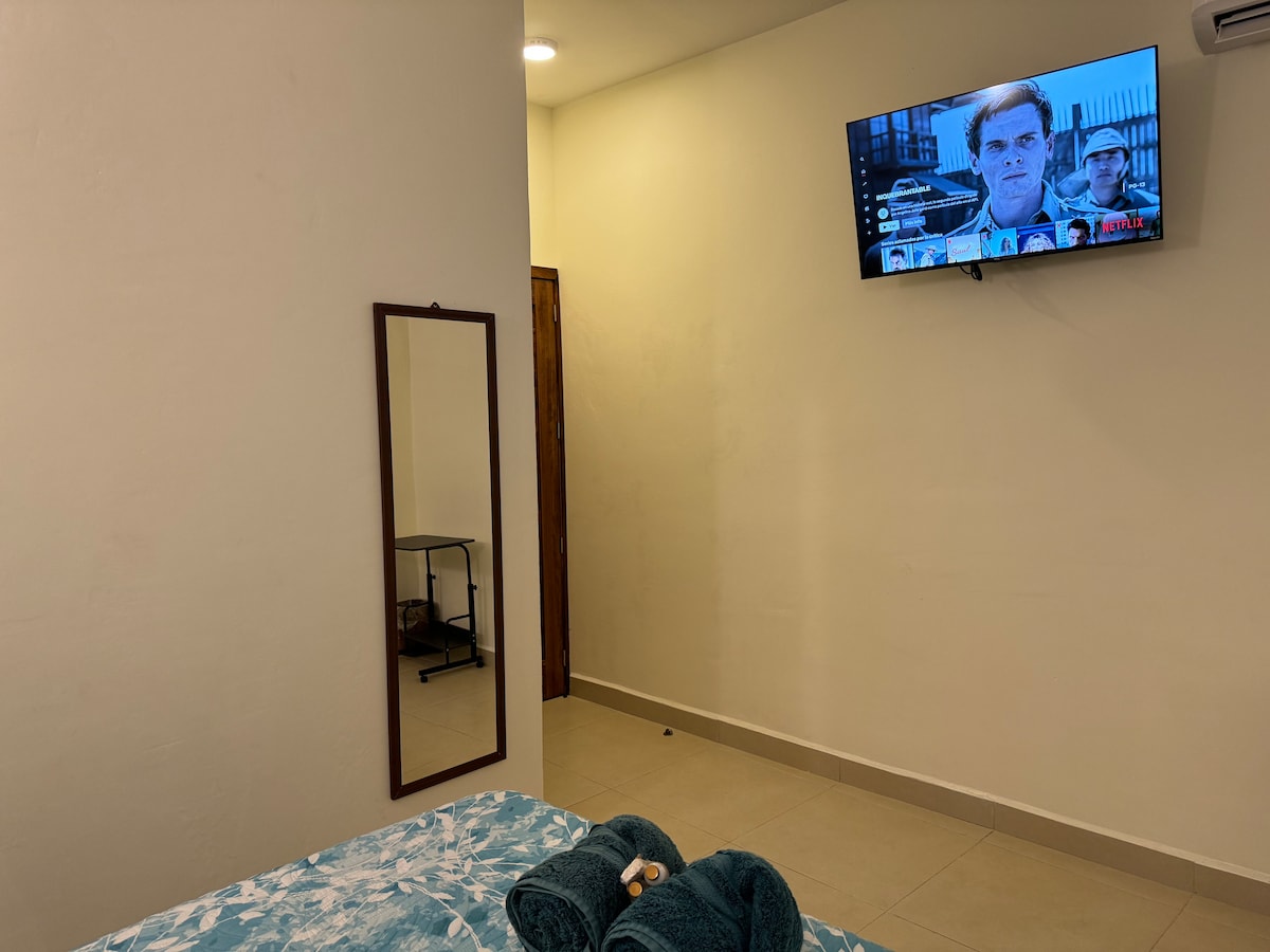 A simple and functional room is depicted, featuring a spacious bed with a blue patterned coverlet. A flat-screen television mounted on the wall is visible, along with a mirror standing in the corner. Soft lighting enhances the neutral-toned walls and tiled floor.