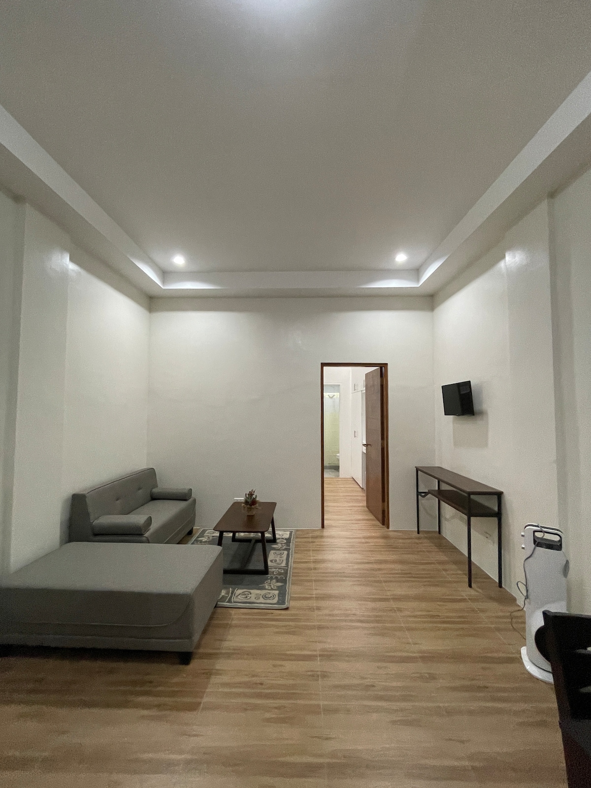 A clean and spacious living area is highlighted by soft lighting and modern furnishings. A gray sofa complements a wooden coffee table, while a narrow console table with a wall-mounted television enhances the room's functionality. Light-colored walls contribute to an open atmosphere.