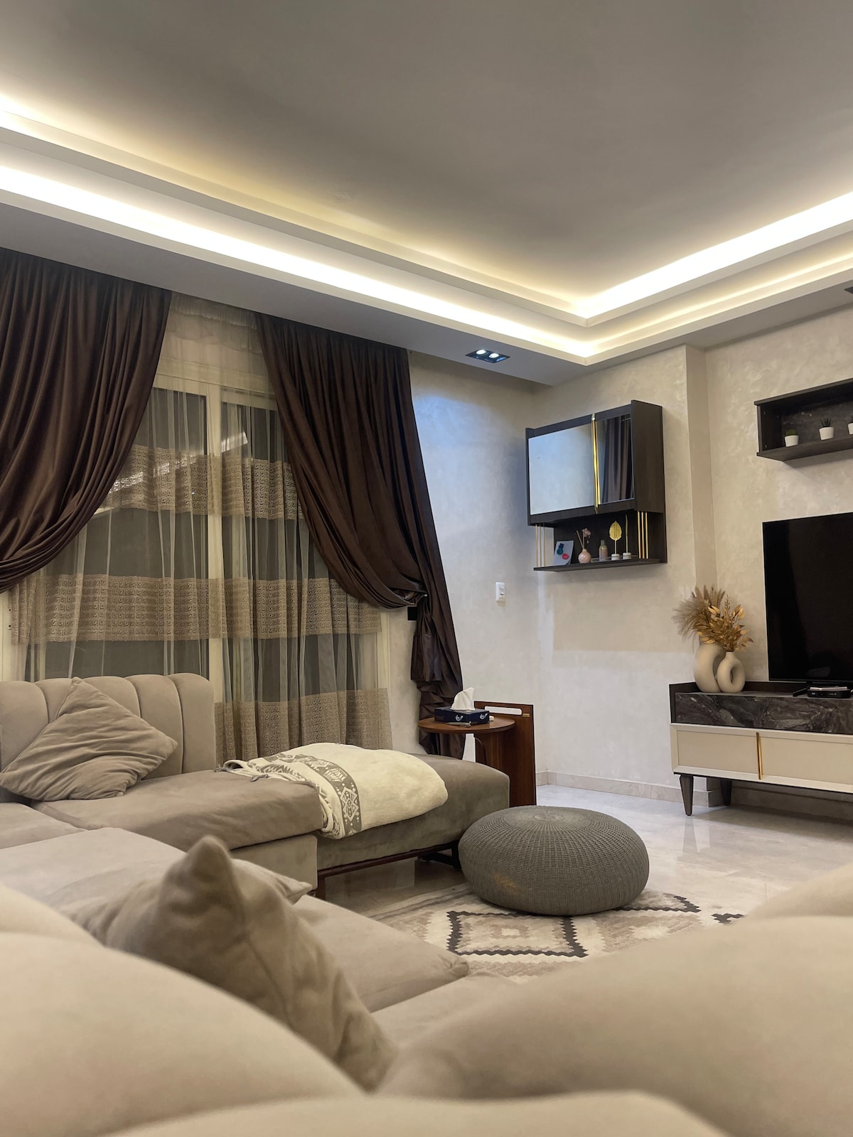 A stylish living room features a soft sectional sofa positioned near a modern television. Elegant curtains frame a large window, and soft lighting accentuates the room's calming palette. A decorative pouf and a textured area rug add layers to the inviting space.