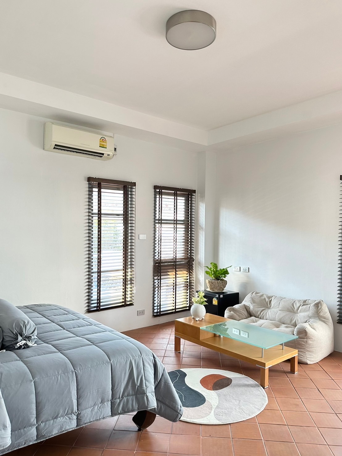 A tranquil bedroom layout features a spacious bed with a gray coverlet. A small glass table is placed beside a plush seating area, complemented by a decorative rug with a subtle design. Natural light filters through the partially open blinds of two windows.